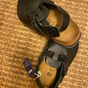 Svarta Birkenstock Boston sandaler - Stilrena svarta Birkenstock Boston sandaler med justerbart spänne och öppen häl. klassisk korkfotbädd för extra komfort. Sulan är i slitstark gummi och har tydlig Birkenstock-logga undertill. Perfekta för en avslappnad och trendig look.