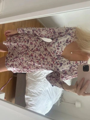 Blommig kortklänning med v-ringning - Supersöt kortklänning med v-ringning och långa ärmar. Klänningen är vit med små lila och rosa blommor, har en lätt volangkant nedtill och är gjord i ett tunt, mjukt material som känns perfekt för vår och sommar.