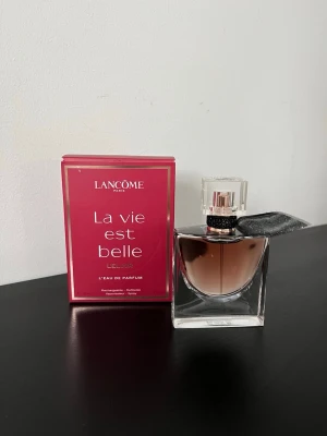 Lancôme La Vie Est Belle L'Elixir parfym - Lancôme La Vie Est Belle L'Elixir är en elegant parfym i en genomskinlig glasflaska med roséguldigt lock och svart dekorband. Doften kommer i en snygg rosa kartong med guldiga detaljer. Flaskan har en modern, fyrkantig form och en exklusiv känsla.