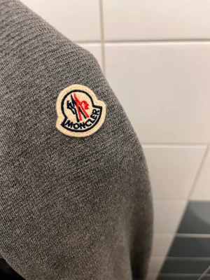 Grå Moncler tröja med patch - Snygg grå tröja från Moncler med ikonisk patch på ärmen. Tröjan har stickad struktur och detaljer i vadderat material på ärmarna, vilket ger en modern och sportig look. Perfekt för dig som gillar exklusiva streetwear-plagg.