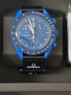 Blå Omega Speedmaster klocka - Snygg blå Omega Speedmaster klocka med svart läderband och vit logga på bandet. Klockan har kronograf, tachymeter och flera urtavlor. Boetten är blå och urtavlan matchar i samma färg. Perfekt accessoar för dig som gillar sportig och modern stil.