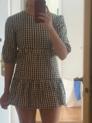 Svartvit rutig miniklänning - Kort klänning med svartvitt rutmönster och puffärmar. Klänningen har rund halsringning och volang nedtill, vilket ger en söt och trendig look. Perfekt för dig som gillar en avslappnad men ändå stilsäker outfit.