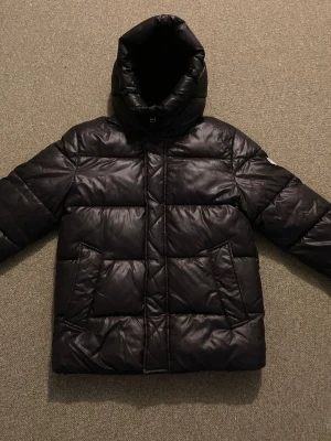 Svart pufferjacka från Michael Kors - Snygg svart pufferjacka från Michael Kors med huva och dragkedja. Jackan har breda, quiltade paneler och två stora fickor framtill. Materialet är glansigt och ger en modern look, perfekt för kalla vinterdagar.