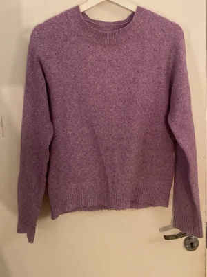 Ljuslila stickad tröja - Mysig ljuslila stickad tröja med rund halsringning och långa ärmar. Tröjan har ribbade muddar vid ärmslut och nederkant, och är tillverkad i ett mjukt material som känns skönt mot huden. Perfekt för lager på lager och enkel att matcha med jeans eller kjol.