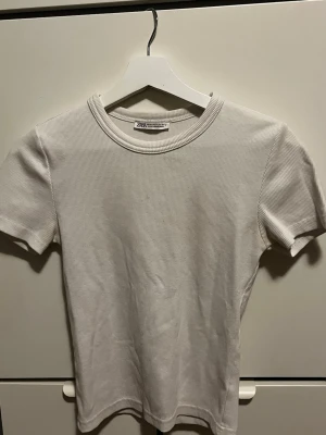 Ribbad vit t-shirt från Zara - Säljer en stilren ribbad t-shirt från Zara i vit färg. T-shirten har korta ärmar, rund halsringning och är tillverkad i ett mjukt bomullsmaterial. Perfekt basplagg till garderoben och enkel att matcha med allt.