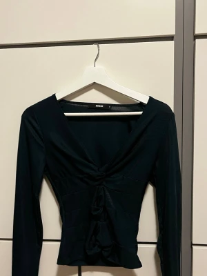 Grön långärmad topp från BikBok - Trendig grön långärmad topp från BikBok med v-ringning och snygga knytningar framtill. Toppen har en ribbad struktur och figurnära passform som ger en cool och modern look. Perfekt att styla med jeans eller kjol för en uppdaterad outfit.