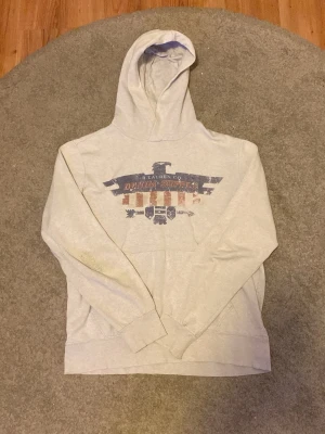 Grå hoodie från Denim & Supply Ralph Lauren - Ljusgrå hoodie med stor huva och tryck framtill i blått och rött med texten 'Denim & Supply R Lauren Co' och en örn. Mjuk bomullskänsla och avslappnad passform, perfekt för en chill look. Tyvärr slitage på högra armbågen som man ser på den första bilden.