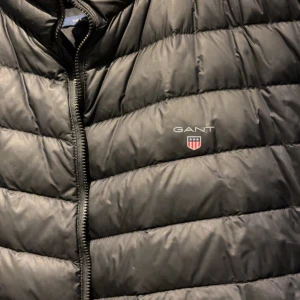 Svart dunjacka från GANT - Snygg svart dunjacka från GANT med quiltad design och diskret logga på bröstet. Jackan har dragkedja framtill och står upp lite i kragen för extra värme. Perfekt för kalla vinterdagar och riktigt stilren look.