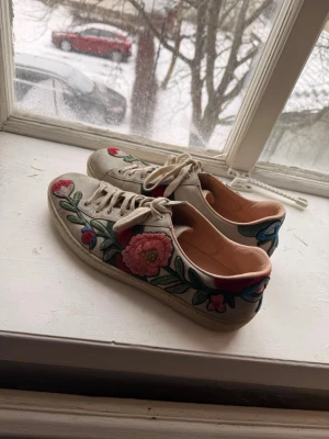 Gucci sneakers med broderade blommor - Snygga sneakers från Gucci i beige läder med broderade blommor i rött, rosa, grönt och blått. Klassisk låg modell med vita snören och ikoniska gröna och röda ränder på sidan. Insidan är ljusrosa och sulan är vit. Perfekta för dig som vill sticka ut med din stil.