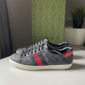 Gucci sneakers med röd och blå detalj - Snygga Gucci sneakers i mörkgrått med klassiska röda och blå ränder på sidan. Skorna har skinnpaneler i rött och blått med ormskinnsstruktur på hälen och vit sula. Diskret GG-mönster i tyget och snörning framtill. Perfekta för dig som vill sticka ut med lyxig streetstyle. Skick 9/10, storlek 42 men kan även passa 43. Pris går att diskutera:)