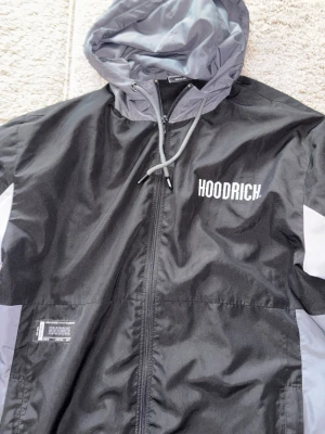 Svart och grå vindjacka från Hoodrich - Snygg vindjacka från Hoodrich i svart och ljusgrått med vita logotexter på bröstet och ryggen. Jackan har huva med dragsko, hel dragkedja framtill och kontrastpaneler på ärmarna. Perfekt för streetwear-stil och skyddar mot vind.