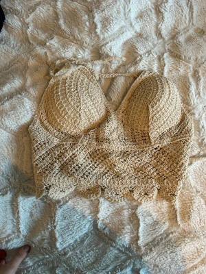 Beige virkad linnetopp med knyt - Superfin beige virkad linnetopp med knyt i nacken och vadderade kupor. Toppen har ett snyggt mönster och är perfekt för varma dagar. Ger en boho-vibe och är lätt att matcha med jeans eller kjol.