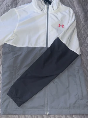 Under Armour vindjacka vit/grå - Snygg vindjacka från Under Armour i vitt och grått med svarta ärmar. Jackan har huva, dragkedja framtill och två sidofickor. Under Armour-logga i rött på bröstet. Perfekt för blåsiga dagar och sportiga outfits.