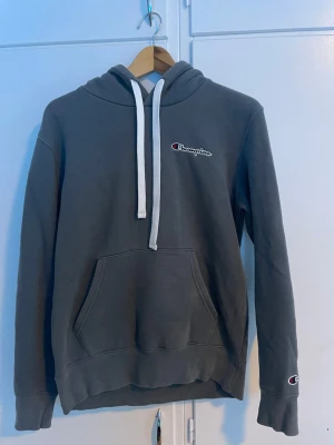 MörkGrå hoodie från Champion - Snygg mörkgrå hoodie från Champion med vit dragsko och klassisk logga på bröstet. Hoodien har en stor magficka och ribbade muddar vid ärmar och nederkant. Perfekt för en avslappnad och sportig stil.