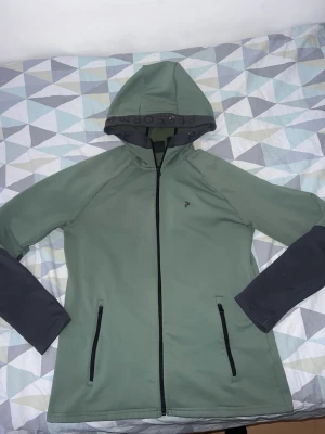 Grön hoodie med svarta detaljer - Snygg grön hoodie från Peak Performance med svarta detaljer på ärmar och dragkedjor. Huva med svart text och två praktiska fickor med dragkedja framtill. Tillverkad i ett mjukt och stretchigt material som passar perfekt till en aktiv livsstil.står att de storlek L men de tight kofta passar som M, kan passa S, har själv S och den sitter bra ! Köpt för 1899 !