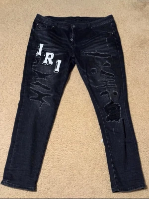 Amiri Jeans  - Unika svarta jeans med raka ben och slitningar framtill. Dekorerade med stora vita bokstavspatchar på både fram- och baksida samt coola distressed detaljer. Jeansen har klassisk femficksmodell och knappgylf. Perfekta för en edgy streetwear-look.