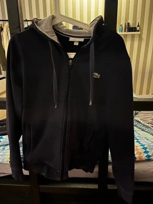 Lacoste zip tröja - Snygg svart hoodie från Lacoste Sport med dragkedja framtill och klassisk krokodil-logga på bröstet. Grå insida på huvan och justerbara snören. Två fickor framtill och ribbade muddar. Perfekt för en chill och sportig look.