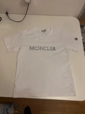 Vit t-shirt med modern logga - Snygg vit t-shirt med stor logotyp tryckt över bröstet och ett märke på ärmen. Klassisk rund hals och korta ärmar. Tillverkad i mjuk bomull för en skön känsla och clean look.