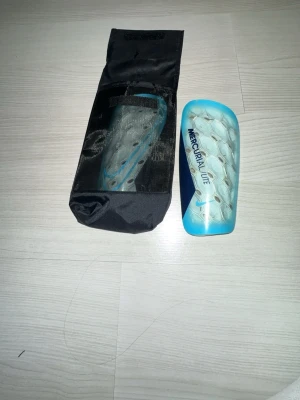 Nike Mercurial Lite benskydd blå - Nike Mercurial Lite benskydd i blått och silver med hexagonmönster och svart Nike-logga. Tillverkade i plast och skum för bra skydd vid fotboll. Kommer med en svart förvaringspåse.