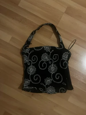 Svart topp med blommigt mönster - Svart linnetopp med vita broderade blommor och virvlande mönster. Toppen har breda axelband och en rak, något croppad passform. Perfekt för dig som gillar unika detaljer och vill sticka ut lite extra. Dragkedja finns på sidan! Storlek S