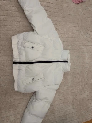 Vit pufferjacka med svarta detaljer - Säljer en vit pufferjacka med svarta detaljer och dragkedja. Jackan har två snedställda fickor med knappstängning och ribbad nederkant. Perfekt för kalla vinterdagar och ger en cool look.