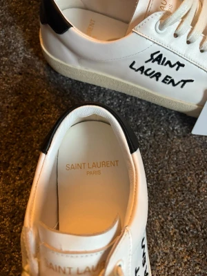 Saint Laurent vita sneakers - Snygga vita sneakers från Saint Laurent med svart häl och handskriven logga på sidan. Skorna har klassisk låg profil, vita snören och sula i gummi. Tillverkade i läder för en lyxig känsla och stilren look.