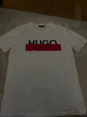 Vit HUGO t-shirt med röd detalj - Vit t-shirt från HUGO med svart logotyp och en bred röd rand över bröstet. Klassisk passform och rund halsringning. Perfekt för en stilren och modern look.