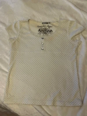 Vit topp med silverprickar Guardian Angels - Kortärmad vit topp från Guardian Angels med små silverprickar över hela tyget och dekorativa knappar framtill.