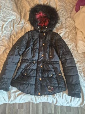 Svart dunjacka med mönster och fuskpäls  - Svart dunjacka från Yes! By CoolCat med snygga svarta spetsmönster, guldfärgade knappar och dragkedja. Jackan har huva med svart fuskpäls och röd insida, samt dragsko i midjan för en figurnära passform.