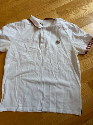 Vit Moncler pikétröja - Klassisk vit pikétröja från Moncler med krage och knäppning framtill. Tröjan har röda och marinblå ränder på kragen och ärmsluten samt Moncler-logga på bröstet. Perfekt till en avslappnad men stilren look.