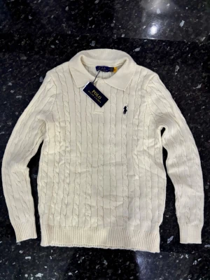 Vit kabelstickad tröja Polo Ralph Lauren - Kabelstickad vit tröja från Polo Ralph Lauren med klassisk krage och broderad logga på bröstet. Tröjan har långa ärmar, ribbade muddar och är tillverkad i mjukt material 