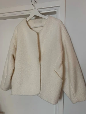 Krämvit oversized teddyjacka - Mysig och oversized teddyjacka i krämvit färg med rundad halsringning och breda ärmar. Jackan har en fluffig, mjuk struktur och minimalistisk design utan synliga knappar eller dragkedja. Perfekt för att hålla värmen under kyliga dagar.