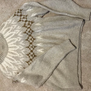 Beige stickad tröja från Lindex - Mysig beige stickad tröja från Lindex med klassiskt nordiskt mönster i vitt och brunt runt oket. Tröjan har rund halsringning, långa ärmar och ribbade muddar. Perfekt för kalla dagar och enkel att matcha med jeans eller kjol.