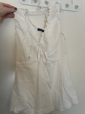 Vit linnetopp med volang och knyt - Söt vit linnetopp från Brandy Melville, med volangkant längs v-ringningen och ett dekorativt knyt framtill. Toppen är ärmlös och har en lätt strukturerad yta som ger en avslappnad känsla. Perfekt för varma dagar och enkel att styla.