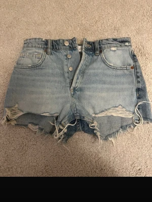 Ljusa denimshorts med slitningar - Snygga ljusblå jeansshorts i denim med medelhög midja, råa kanter och slitna detaljer både fram och bak. Klassisk femficksmodell med knäppning framtill. Perfekta för en avslappnad och trendig sommarstil.