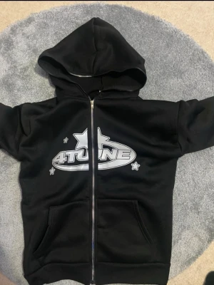 Svart hoodie med 4TUNE-tryck - Svart hoodie med dragkedja och stor huva. Framsidan har ett stort silverfärgat 4TUNE-tryck med stjärnor. Två fickor framtill och ribbade muddar vid ärmslut och nederkant. Perfekt för en avslappnad streetwear-look.