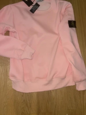 Ljusrosa hoodie från Stone Island - Snygg ljusrosa hoodie från Stone Island med klassisk logopatch på ärmen. Hoodien har rund halsringning, ribbade muddar och är långärmad. Perfekt för dig som vill ha en stilren och trendig look.