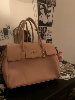 Baby Pink handväska med gulddetaljer - Säljer en stilren beige handväska med två rejäla handtag och lock med guldfärgad vridknapp samt hängande lås. Väskan har en klassisk, boxig form och är tillverkad i slätt konstläder. Perfekt för dig som gillar minimalistisk design och snygga detaljer.