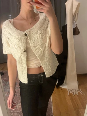 Vit kortärmad stickad kofta - Kortärmad, chunky stickad kofta i offwhite med stora flätstickade detaljer och två knappar framtill. Koftan har en bred krage och är gjord i ett mjukt, papperstjockt garn som ger testad  vintagekänsla. Perfekt att slänga över ett linne! Bra skick! 🥰