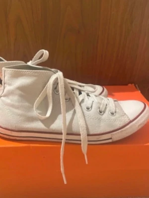 Vita höga Converse sneakers - Klassiska vita höga Converse sneakers i canvas med vita snören och gummisula. Skorna har den ikoniska röda och svarta randen runt sulan och metallöljetter för snörningen. Perfekta för en avslappnad och trendig stil.