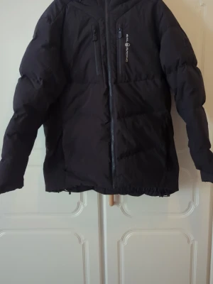 Svart dunjacka från Sail Racing - Svart dunjacka från Sail Racing med huva och två bröstfickor med dragkedja. Jackan har ett stilrent, sportigt snitt och logotyp på bröstet. Perfekt för kalla vinterdagar, tillverkad i ett tåligt material som håller värmen. Köpt på : https://www.booztlet.com/se/sv/sail-racing/patrol-down-jacket_31689604/225650391?volume=5&st=3199&gad_source=1&gad_campaignid=23168119861&gbraid=0AAAAAC3sHKb2JkQpItcLokSbXrwbOig9T&gclid=Cj0KCQiAqeDMBhDcARIsAJEbU9QVUCePUrjPjOTqALQCHzZBd_QdYPObW9I57wJq1RWpDqCyu_gHhpgaA