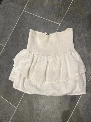 Vit volang tubtopp i linne från BikBok - Söt vit tubtopp med bred smock upptill och dubbla volanger nedtill. Toppen är ärmlös och har en luftig känsla, perfekt för varma dagar. Tillverkad i linne för en naturlig och sval look. Kan användas som kjol också.