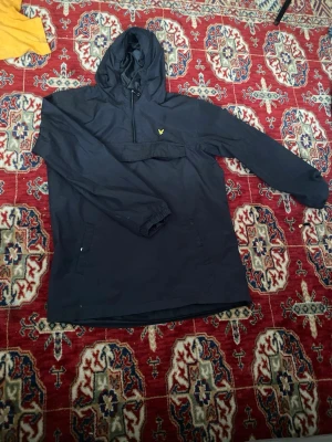 Svart vindjacka från Lyle & Scott - Svart vindjacka från Lyle & Scott med huva och dragkedja vid halsen. Jackan har elastiska muddar vid ärmsluten och en gul logotyp på bröstet. Perfekt för blåsiga dagar och enkel att matcha med olika outfits.