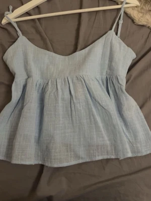 Ljusblå linnetopp med knyt i ryggen - Supersöt ljusblå linnetopp med tunna axelband och vacker knytning i ryggen. Toppen har ett lätt rynkat fall och är tillverkad i ett luftigt material som passar perfekt till varma dagar. Stilren och enkel att matcha med jeans eller kjol.
