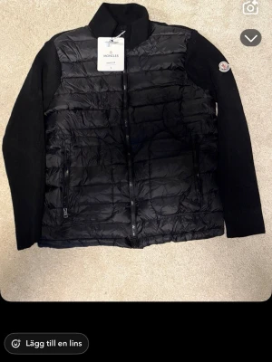 Svart dunväst från Moncler - Snygg svart dunväst från Moncler med stickade svarta ärmar och hög krage. Västen har två dragkedjeförsedda fickor framtill och ikonisk Moncler-logga på ärmen. Materialet är en kombination av dun och stickat tyg för en modern look.den är rätt så liten i storleken så rekommenderar att man bör s-m om du ska köpa den!