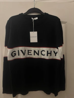 Svart stickad tröja från Givenchy - Snygg svart stickad tröja från Givenchy med vit panel och röd detalj över bröstet där loggan är broderad. Tröjan har rund halsringning och långa ärmar. Perfekt för dig som gillar exklusiva märken och stilren design.