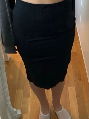 Svart ribbad kjol i midilängd - Säljer en svart, ribbad kjol med kroppsnära passform och hög midja. Kjolen har en enkel och stilren design som passar till många olika looks. Materialet är stretchigt och bekvämt, perfekt för dig som gillar en tight siluett.