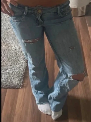 Blå slitna jeans från Il Dolce - Säljer ett par ljusblå jeans från Il Dolce med slitningar och hål på benen för en trendig look. Jeansen har låg midja, raka ben och klassiska fem fickor. Tillverkade i denim med snygga detaljer och blommigt innerfoder i linningen.