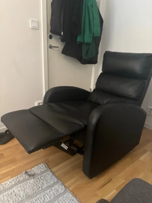 Svart recliner fåtölj i läder - Säljer en svart recliner fåtölj i läder med mjuk stoppning och högt ryggstöd. Fåtöljen har breda armstöd och utfällbart fotstöd för extra komfort. Perfekt för att chilla framför tv:n eller läsa en bok.