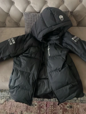 Svart pufferjacka från Sail Racing - Säljer en svart pufferjacka från Sail Racing med huva och dragkedja. Jackan har logga på bröstet och ärmarna samt stora fickor framtill. Tillverkad i slitstarkt material och perfekt för kalla vinterdagar. Snygg och stilren design som håller dig varm.